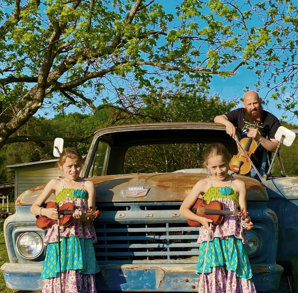 Fiddlin Fluds posing around one of the original, somewhat rusted Ford trucks in a country area during blue skies.