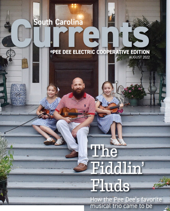 Front cover page of South Carlina Currents magazine, August 2022. Photo of Fiddlin Fluds John, Madelyn Rose, and Caroline sitting on front porch wooden stairs holding on to their fiddles with greenery and plants displayed around them.